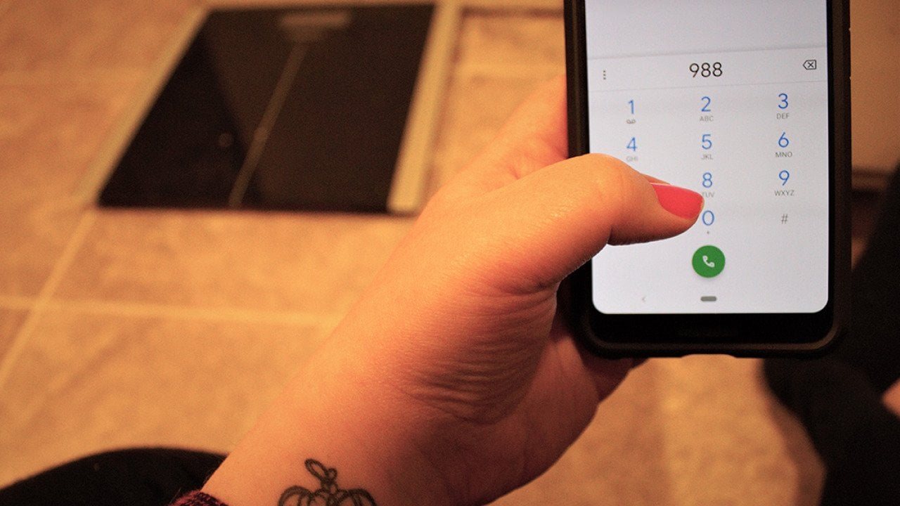 A woman with a tattoo dials the national suicide hotline '988' number from the floor in front of a body weight scale. 
