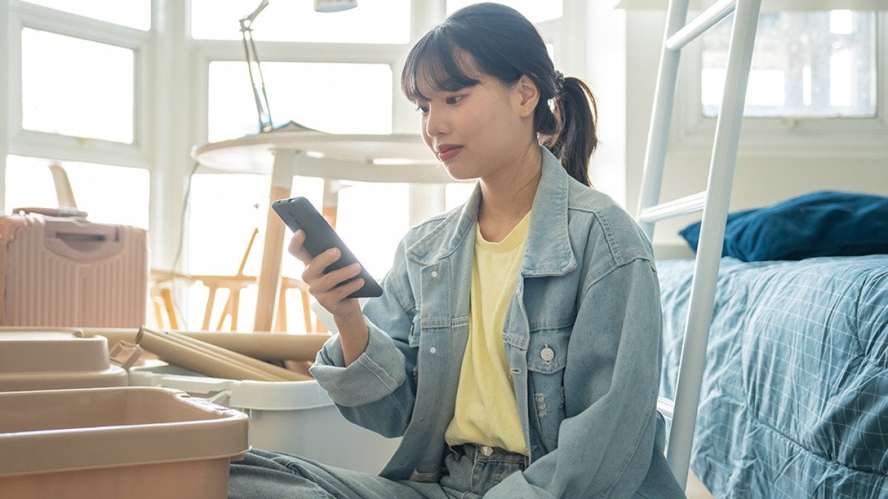 A college student using her smartphone in her dorm while unpacking personal items.