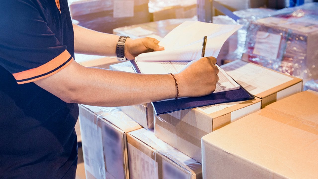 Warehouse worker writing on a clipboard on top of boxes as part of checking inventory.