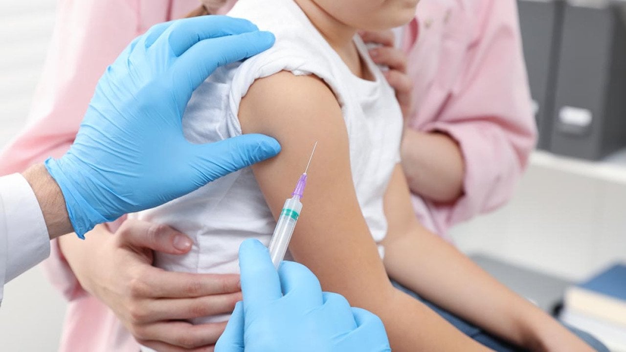 closeup of child's arm with vaccine needle held next to it by a person with blue gloves, an adult in a pink shirt holds child from other side.