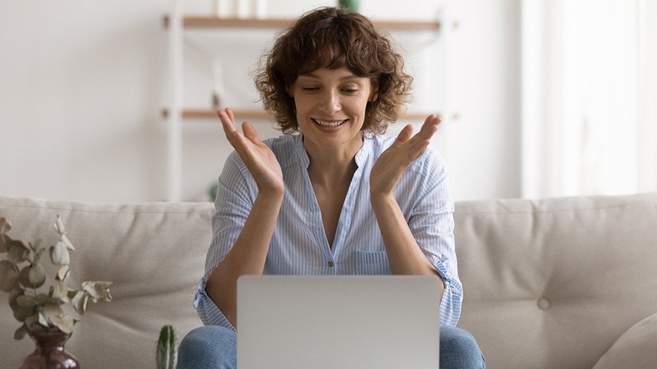A happy woman sitting on her couch reading good news on her laptop.