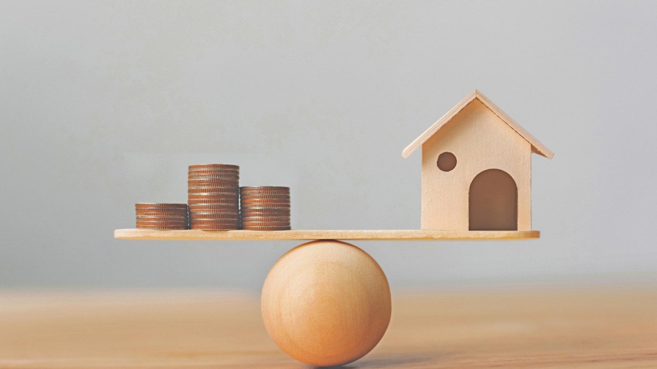 Wooden scale balancing a mini wooden figure of a house on the right and stack of coins on the left.