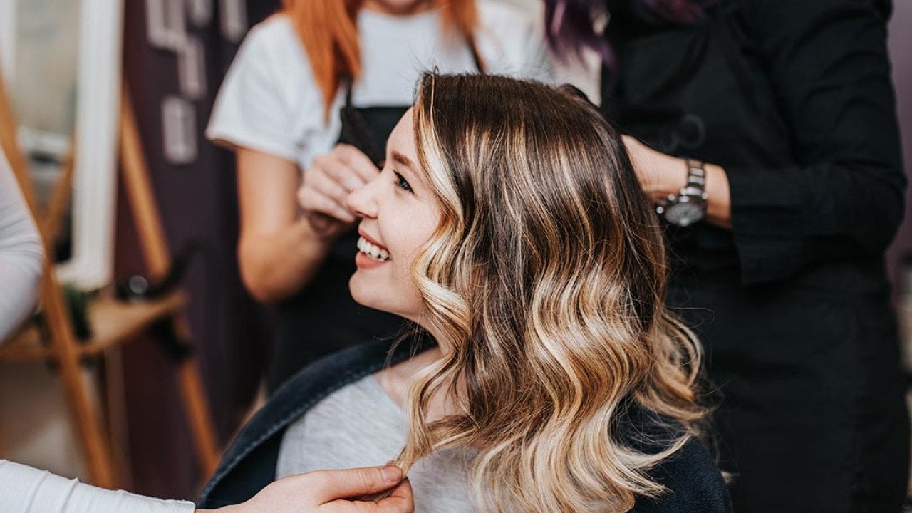 Side profile of woman with blond and brunette hair in long layers smiling at hair salon with two stylists behind her in the background.