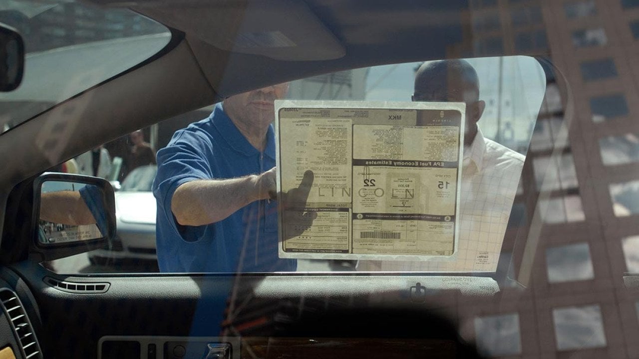 Interior view of car from driver's side looking out passenger window where two people are reading a sticker on the window with the car's features. 