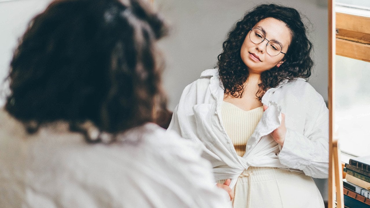 A curvy woman stands in front of a mirror, styling her outfit with a long sleeve shirt.