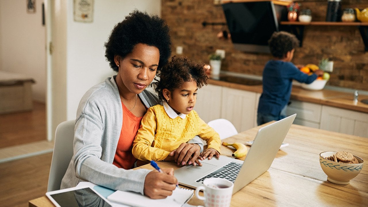 A black mother takes notes as she works at home while her young daughter sits on her lap.