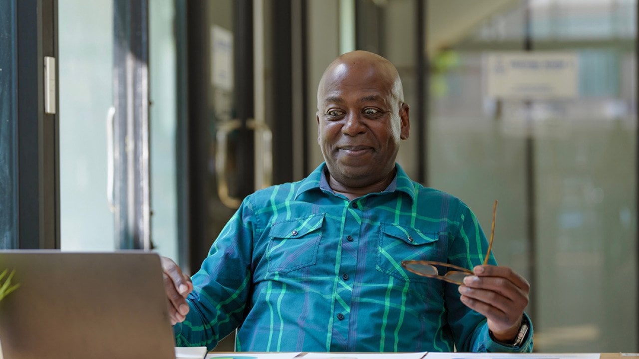 A senior man excitingly looks at financial documents while using a laptop.