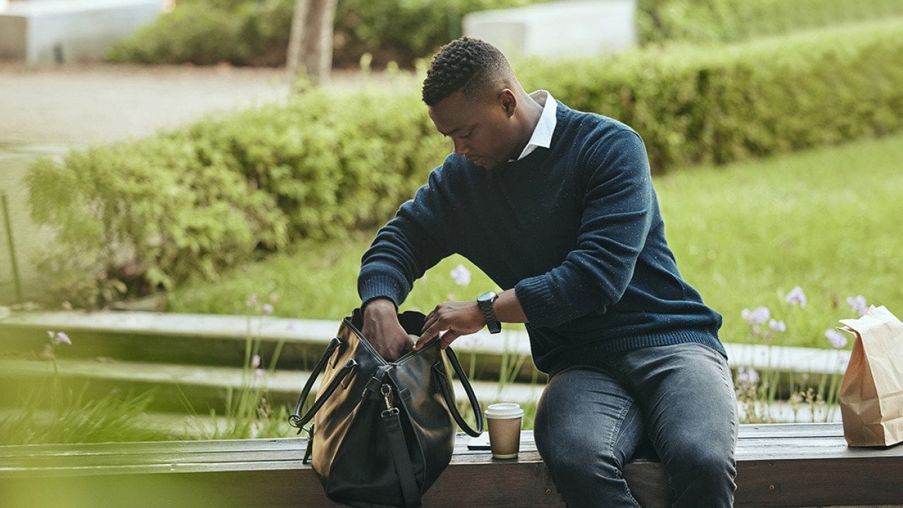 A young black businessman, looking inside his bag to eat lunch while sitting on a bench at a park.