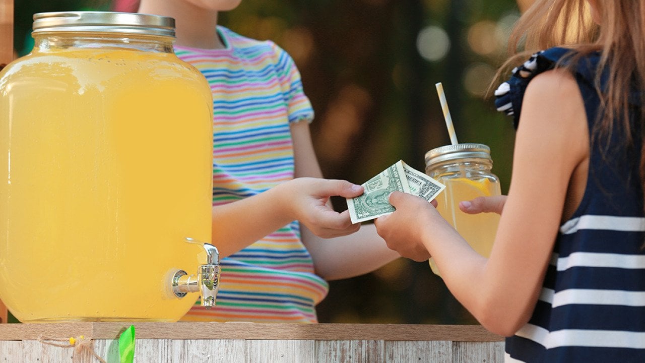 A young girl sells lemonade to another young girl at the park.