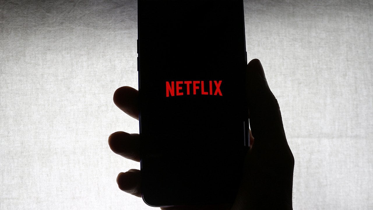 The Netflix logo on the screen of a mobile phone held by a person against a white background.