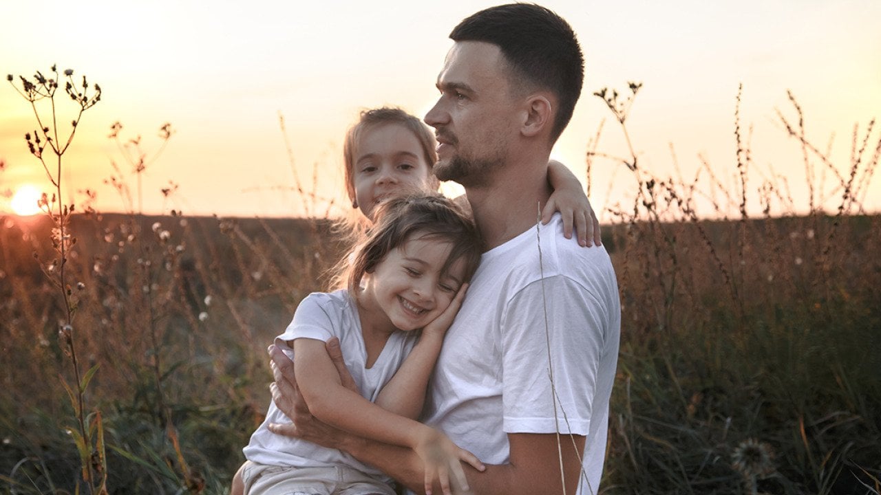 A father with her two young daughters out on a grass field during sunset.