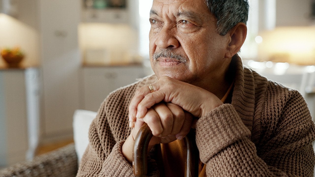 A senior man with a cane thinks deeply while sitting on a sofa.
