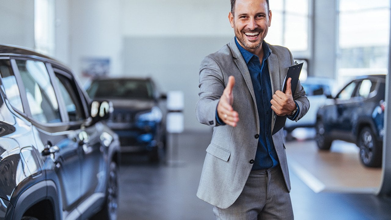 A friendly and cheerful salesman poses to greet car showroom clients with a handshake.