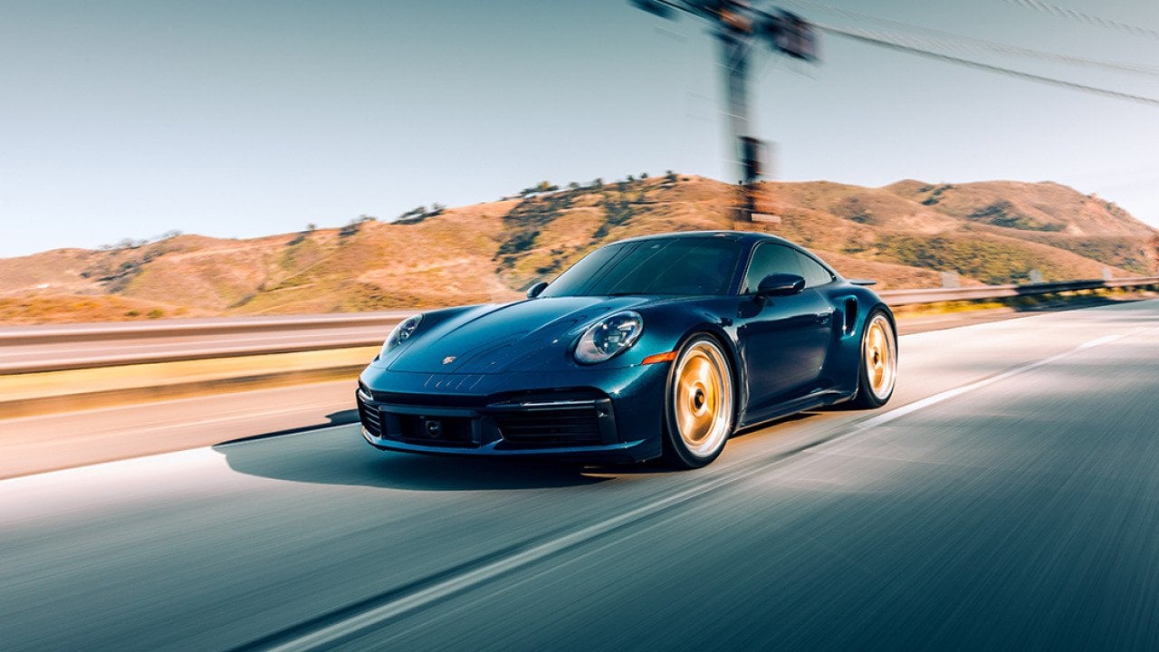 A black Porsche Turbo S driving on a highway with mountains in the background.