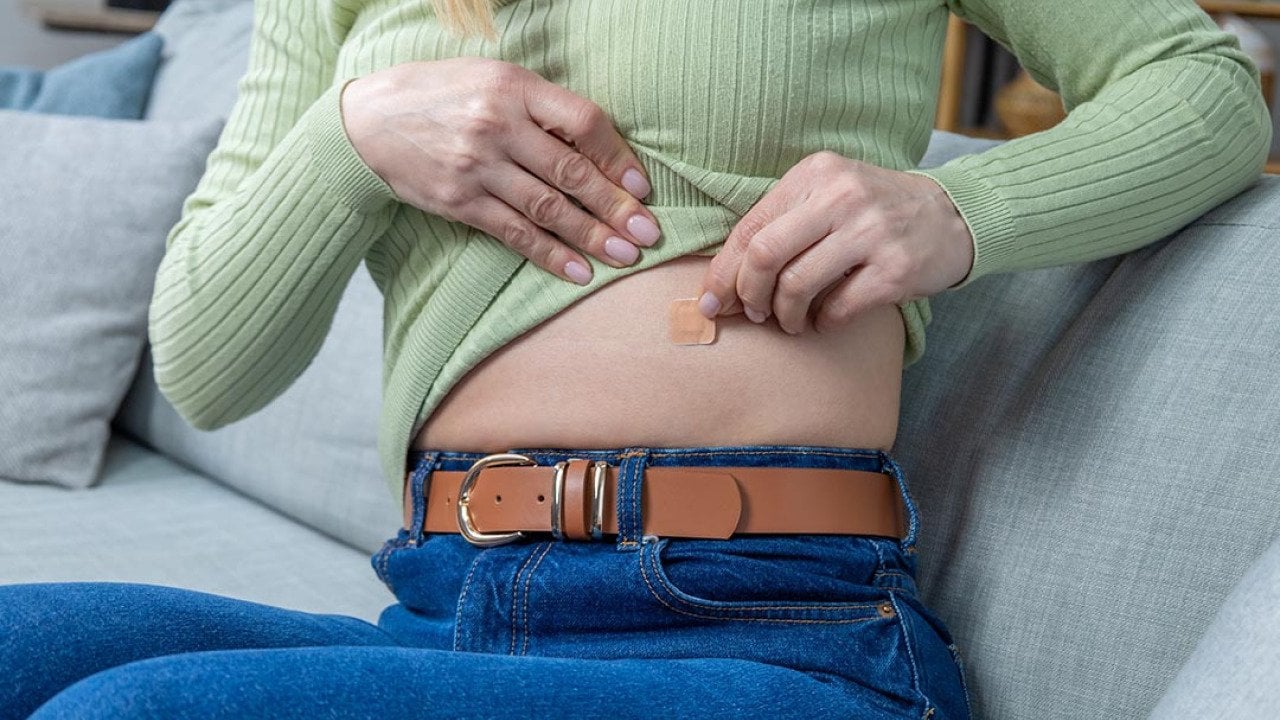 Close up of woman sitting on couch pulling up long sleeve light green shirt to apply hormone replacement patch to her abdomen.
