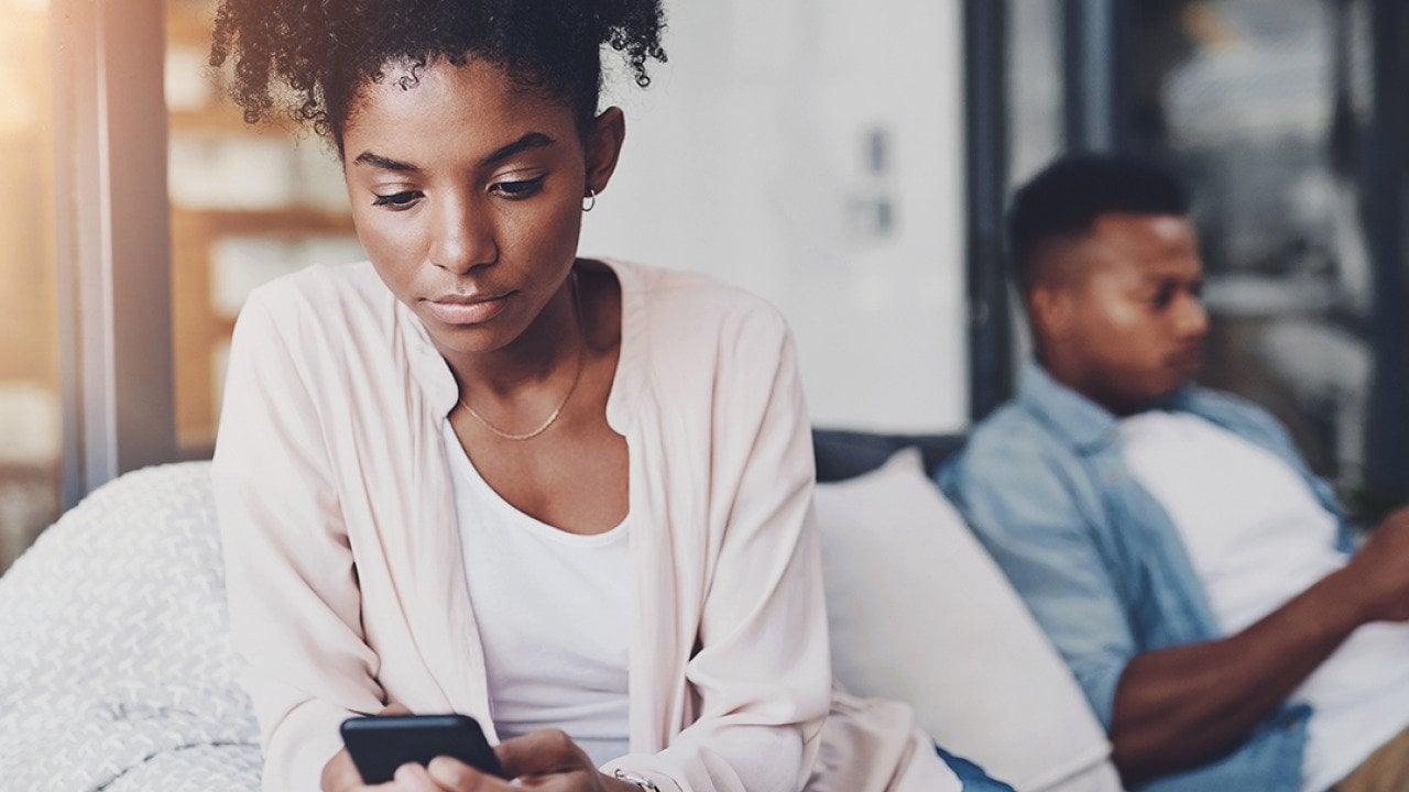 A black couple on their individual phones while sitting apart on the same couch at home.