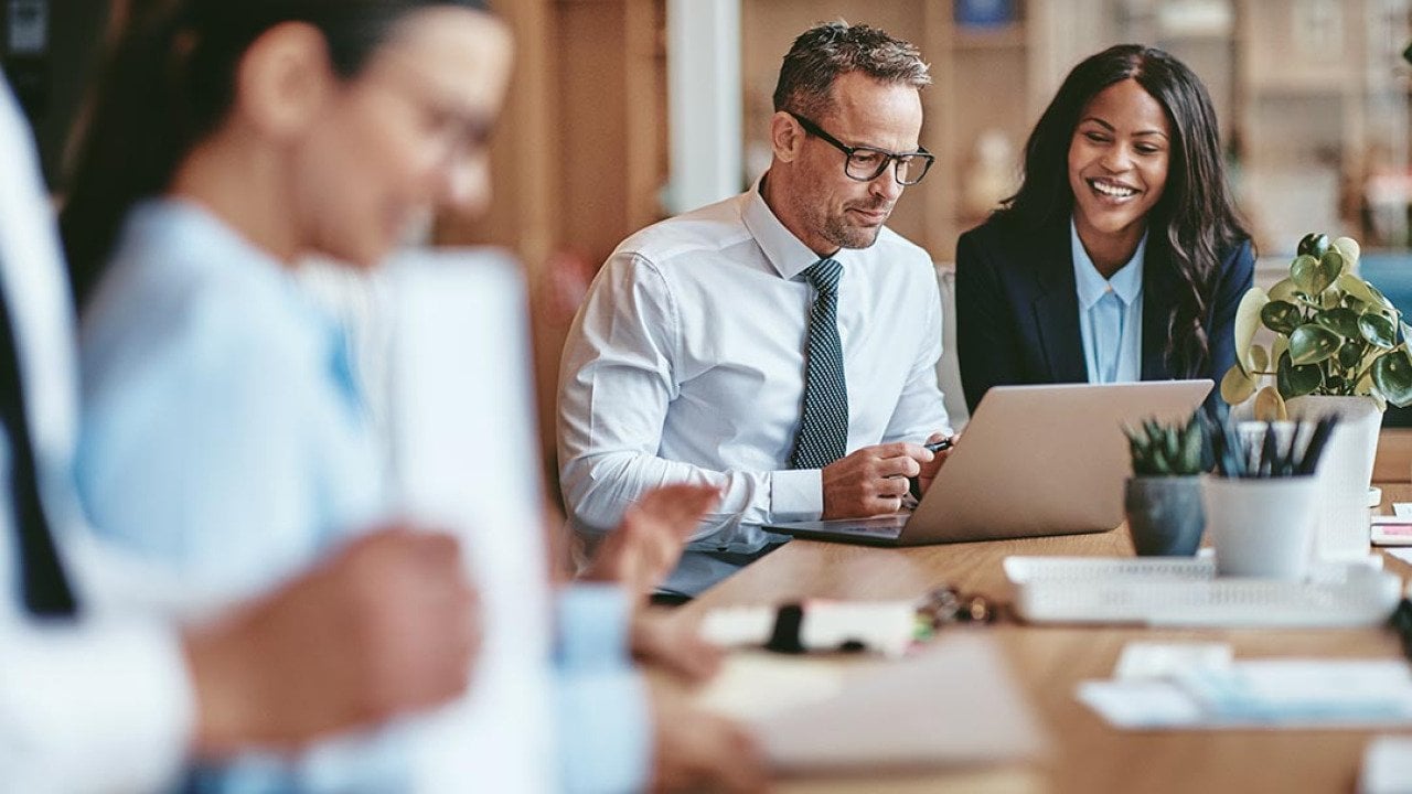 Two diverse businesspeople smiling while working on a laptop together at the end of a boardroom table in an office.