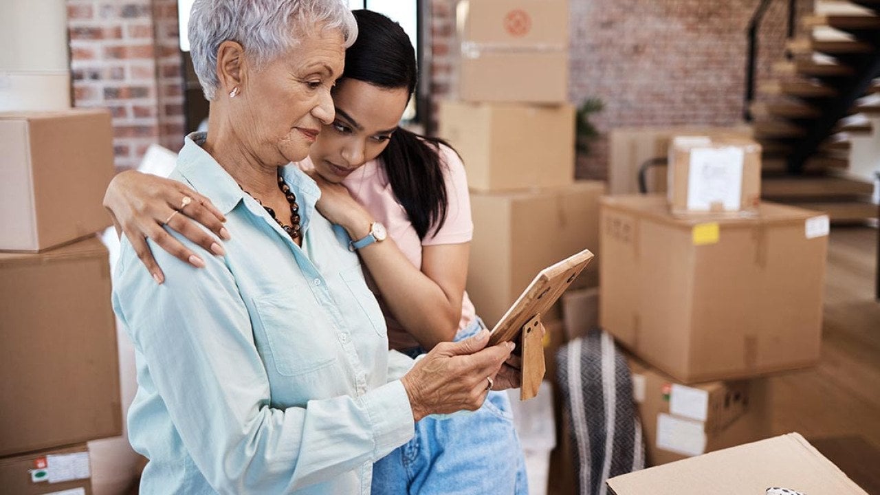 An older woman looking at a photograph while getting a hug from a younger family member while packing boxes on moving day.