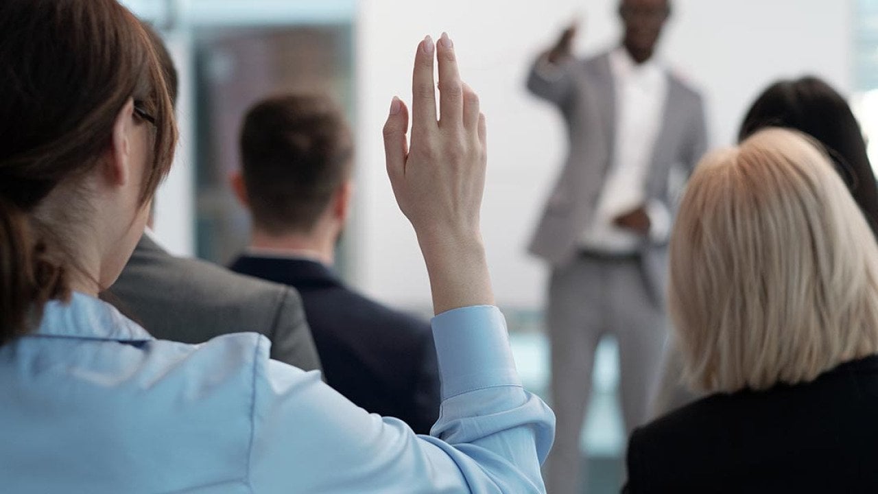 Rear view of raised hand with pointy nails of young employee wishing to answer question of lecturer while sitting behind group of other speakers at conference.