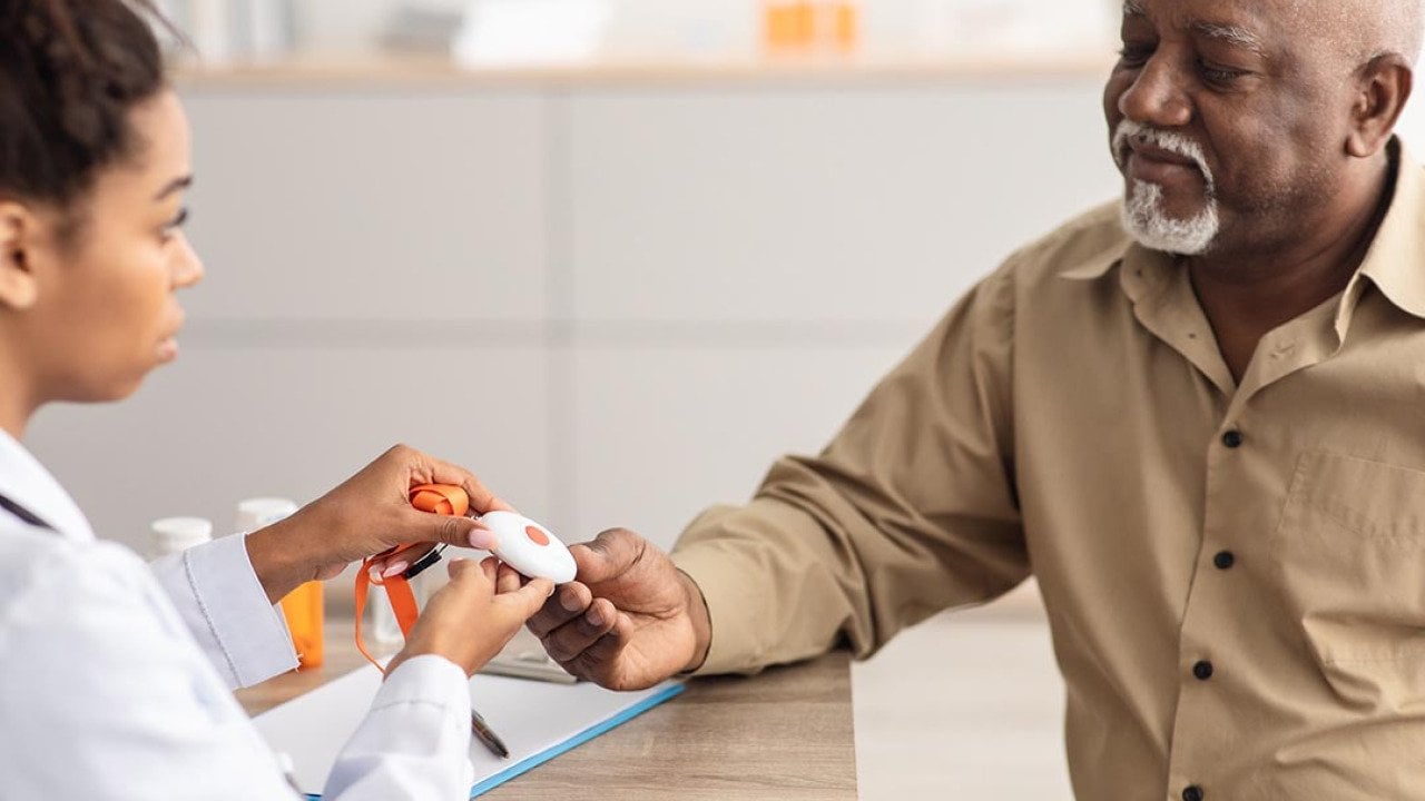 Portrait of black medical worker holding personal alarm button and giving it to mature patient, sitting at table in office, explaining how to use medical alert.