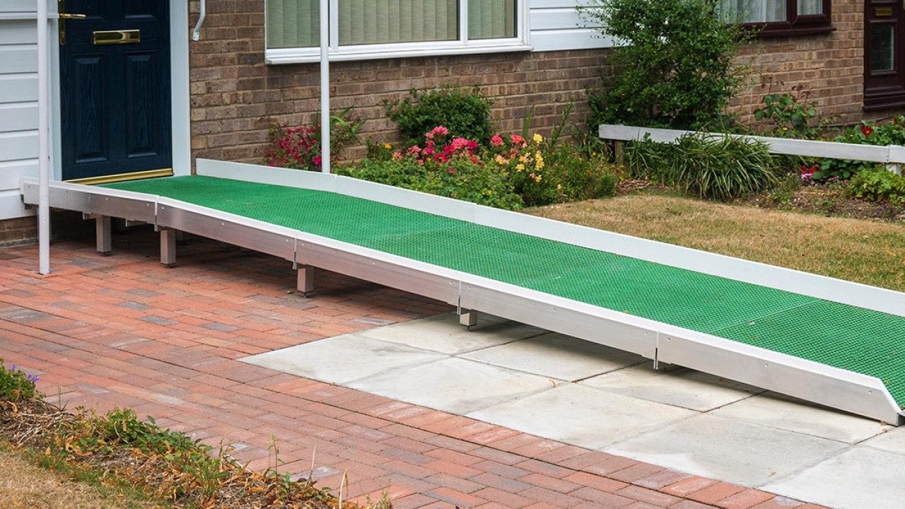 A wheelchair access ramp with green plastic surface installed at the door to a building that has a single step up to a black door.