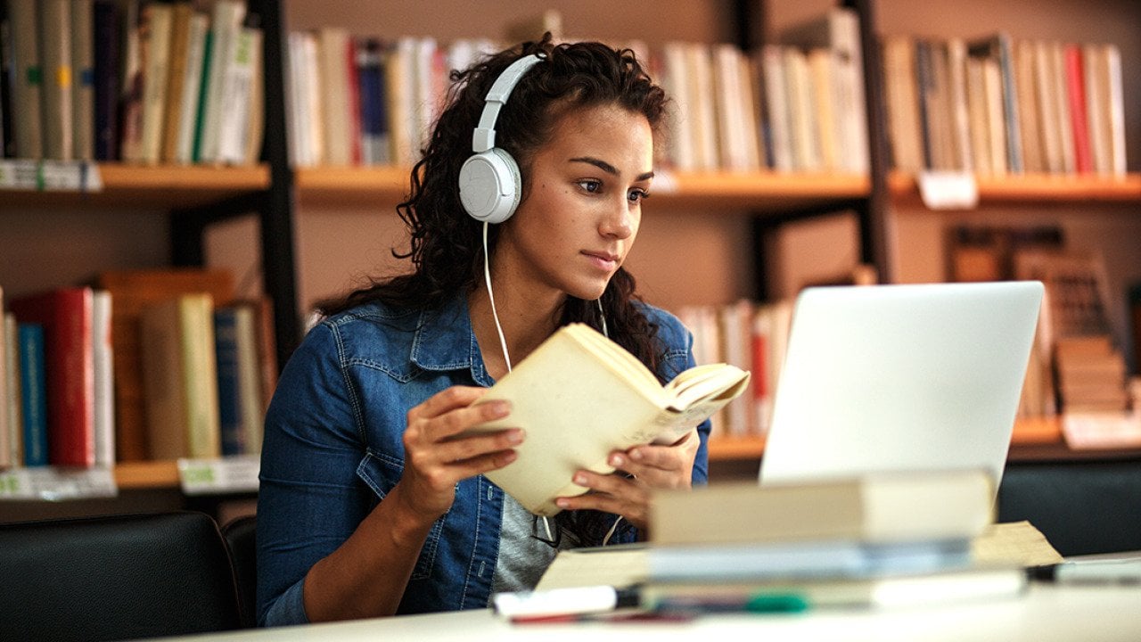 A fema collge student reading a book in a library while listening to music.