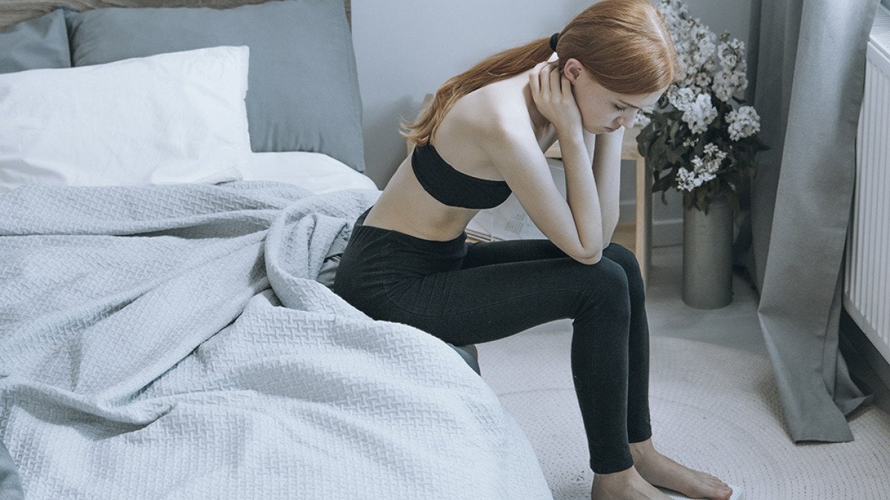 A young woman sits by her bed while pensively checking weight on a scale.
