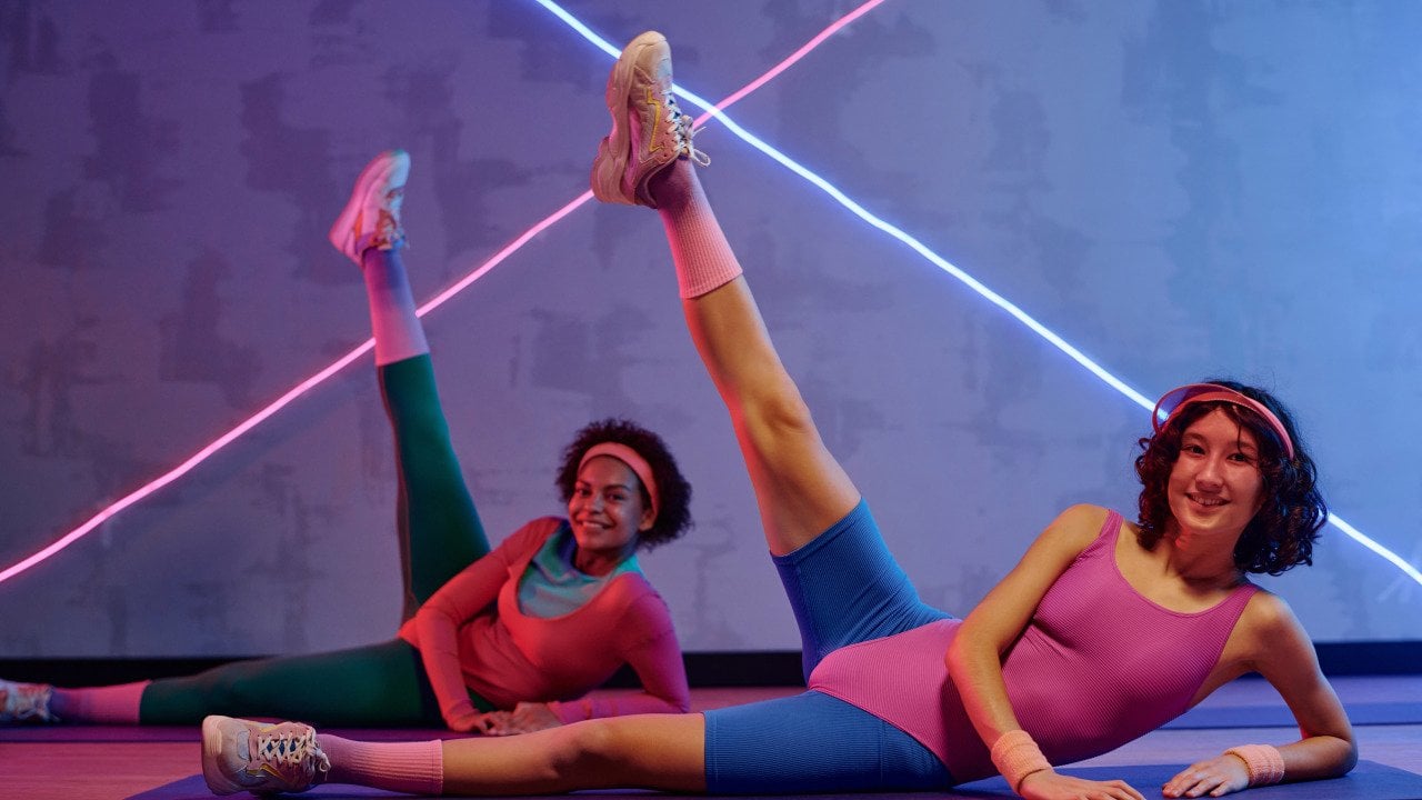 Two young adults wearing 80s style fitness clothing lay on their sides doing side leg raise exercise on floor at gym.