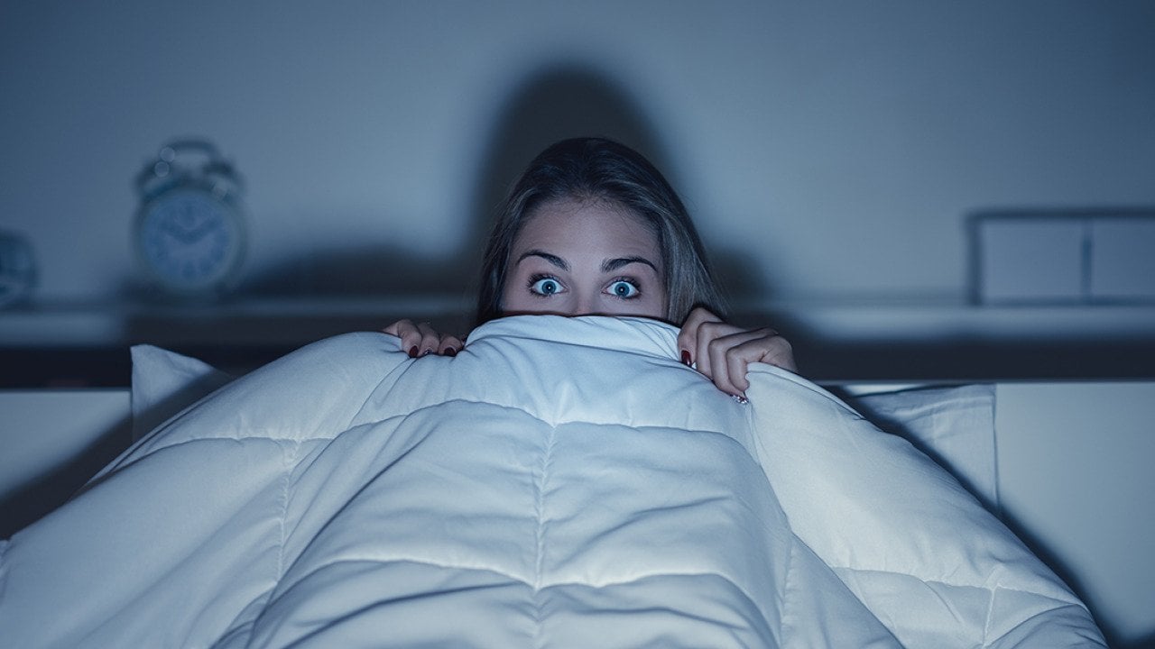 Stylized photo of woman hiding under covers with wide eyes, illuminated by screen from TV.