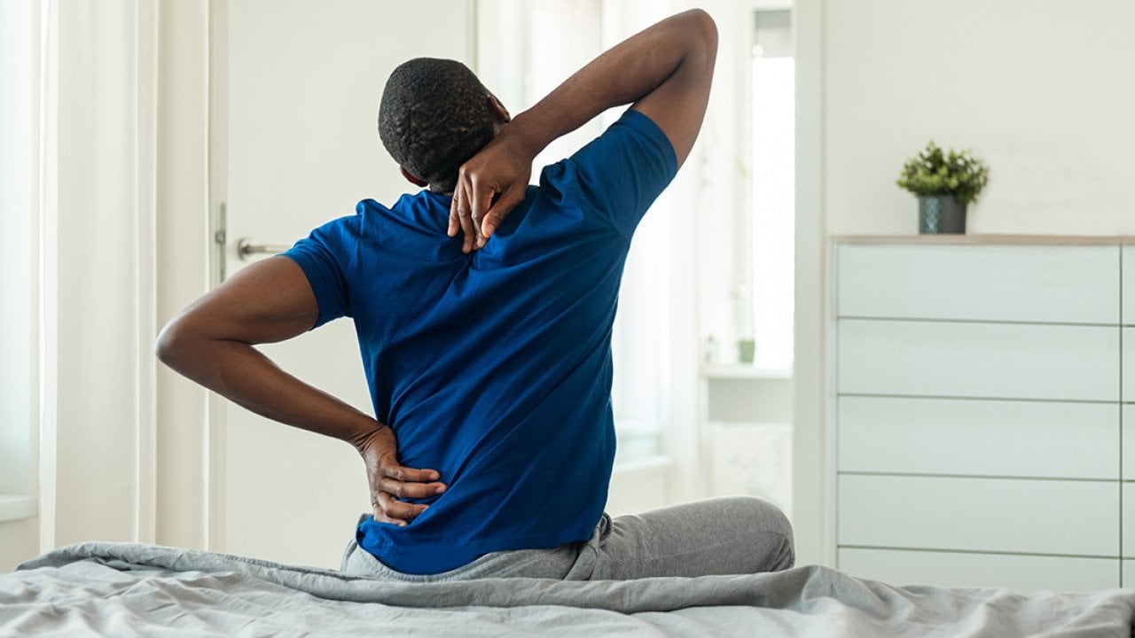 A young black man stretches and reaches for his back after getting up from bed.