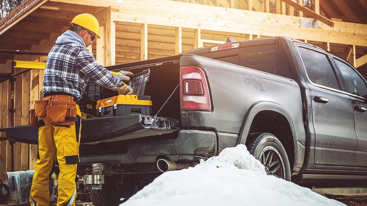 A construction worker in yellow gear prepares tools from a truck at a snowy building site with wooden building framing in the background.