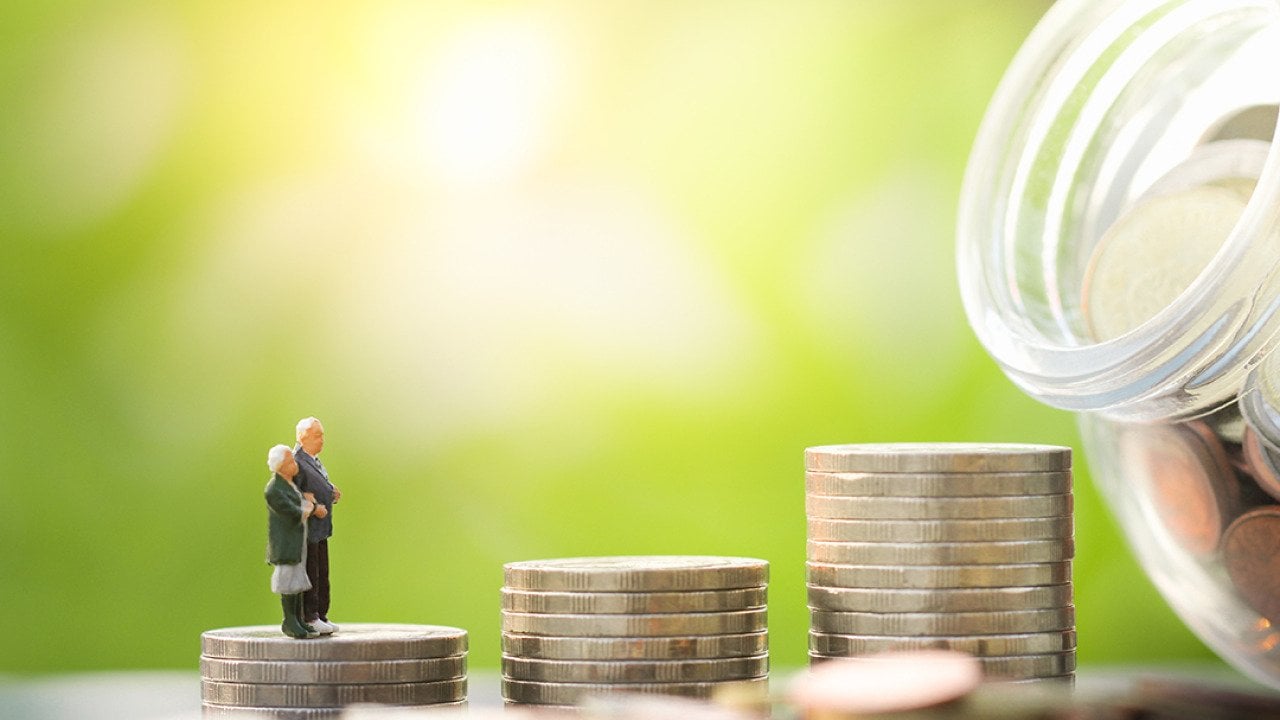 A miniature elderly couple stands on a stack of coins leading to a glass jar full of savings.