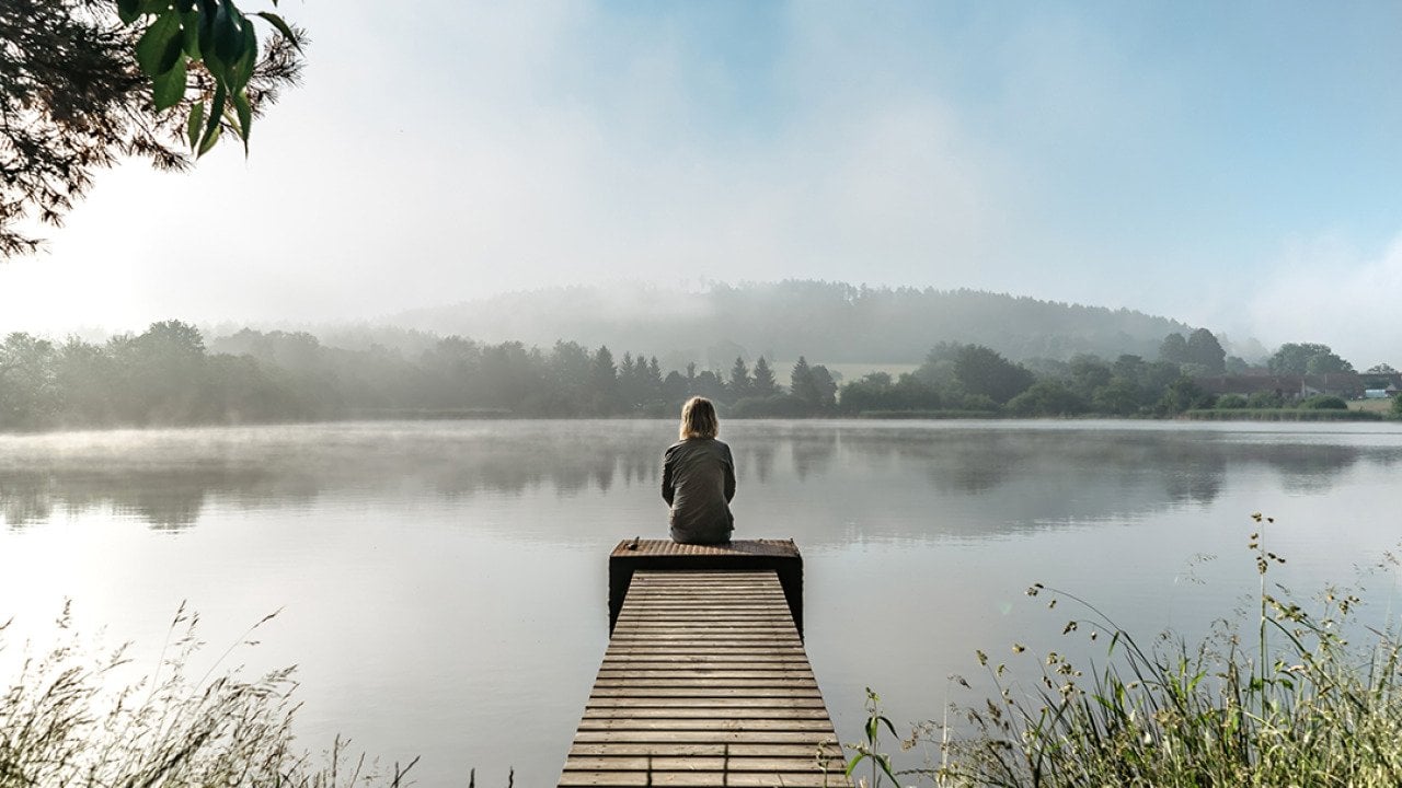 A woman sits quietly at the end of a small pier by a lake during a calm, misty morning.