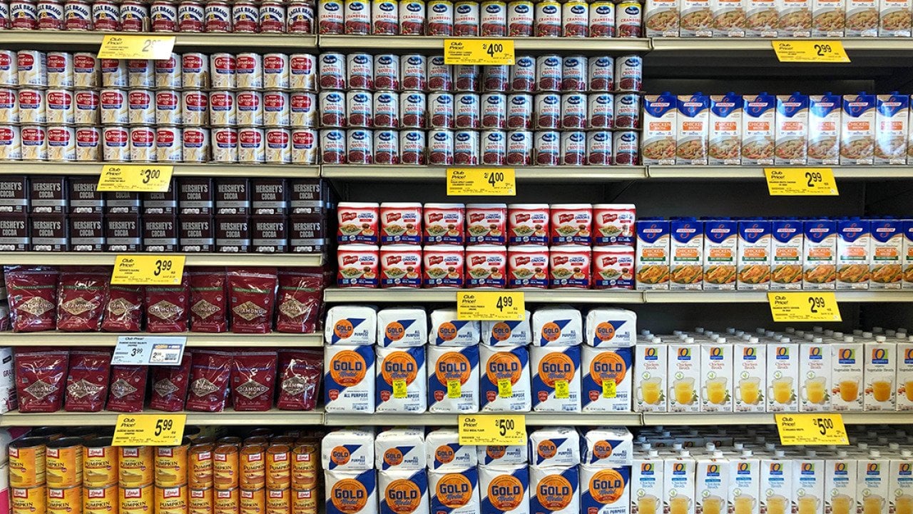 Grocery store shelves of common Thanksgiving and Christmas dinner supplies such as cans of condensed milk, pumpkin puree, and cranberry sauce.