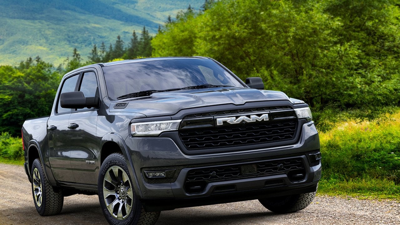 Black 2026 Ram 1500 REV Bighorn driving up dirt road with rolling green hills in background.