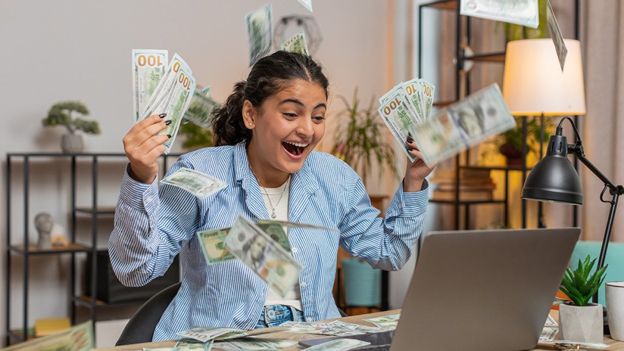 Young person throwing money and bills in the air in front of a computer; concept of getting rich quickly myth