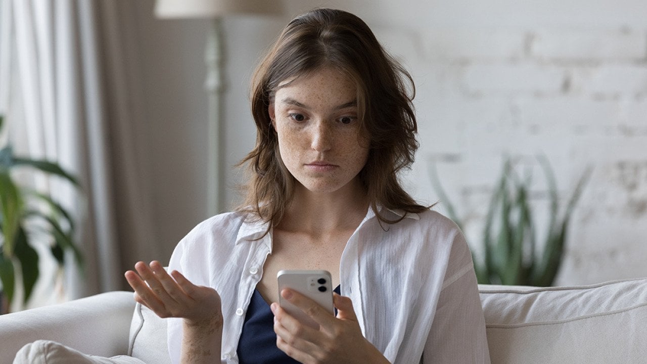 A woman sitting on a couch at home looking concerned at her phone.