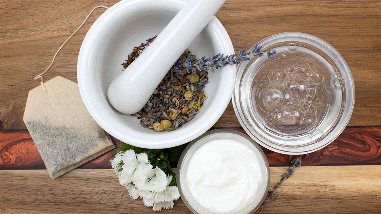 A top view of natural beauty ingredients on a table such as a tea bag, face cream and soap, and a mortar and pestle with dried flowers.