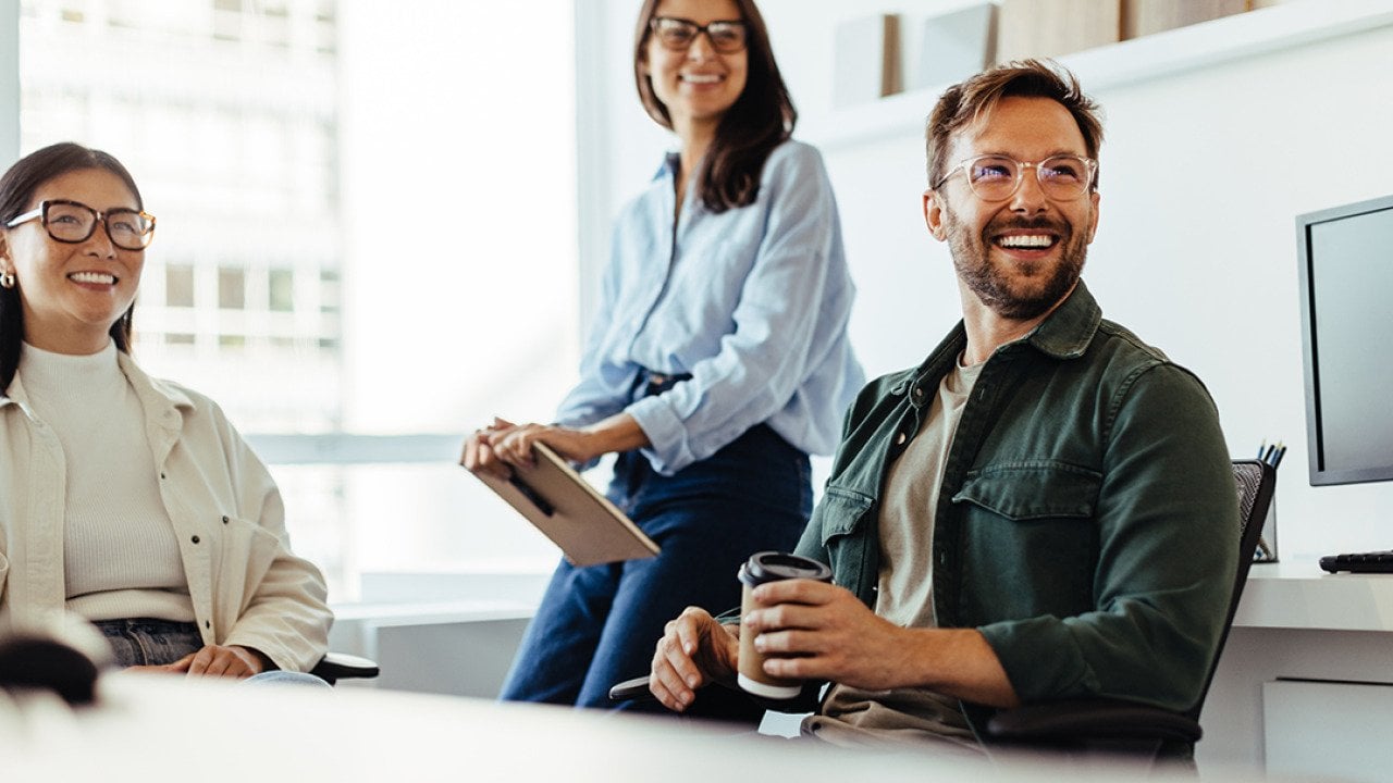 A group of three business people happily listens to a speaker during an office meeting.