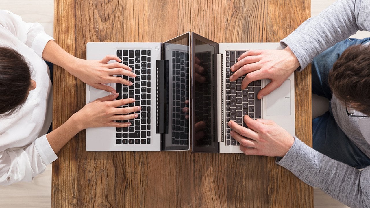 An overhead view of two people using laptops and sitting across each other on a table.