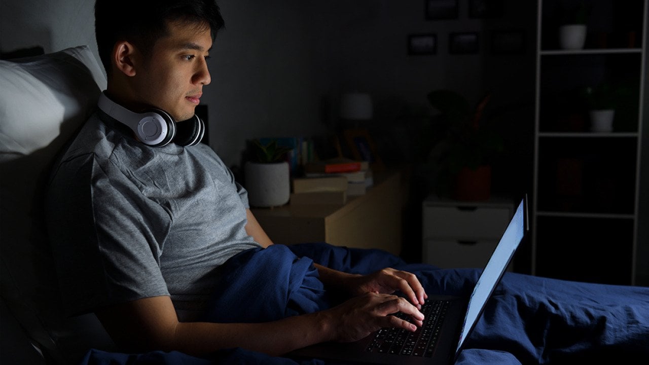 A young man uses his laptop in bed at night.
