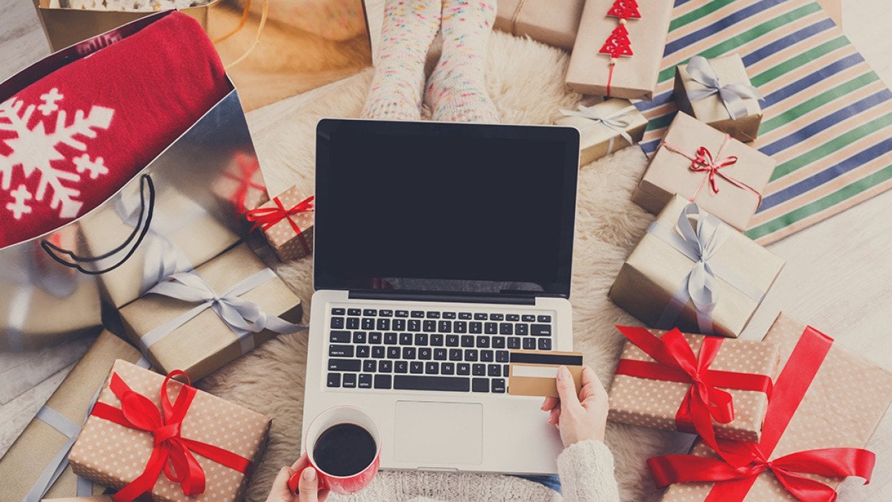 A shopper uses a laptop and credit card while sitting on a bed surrounded by Christmas gifts.