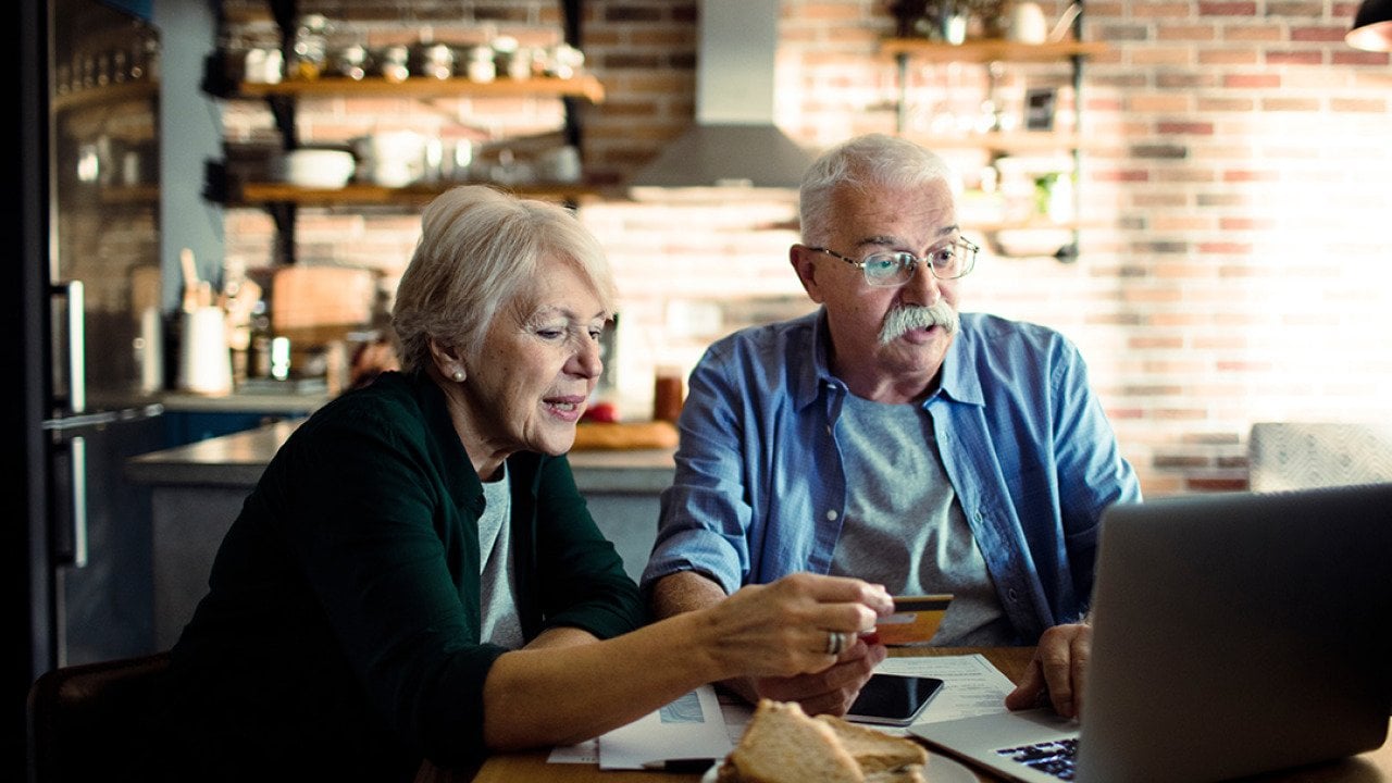 An elderly couple using a credit card at home with a laptop.