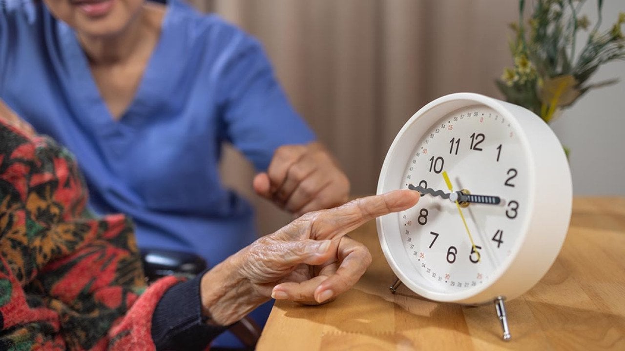 Close up of Asian elderly woman's hand pointing at analog clock with caregiver visible behind her.