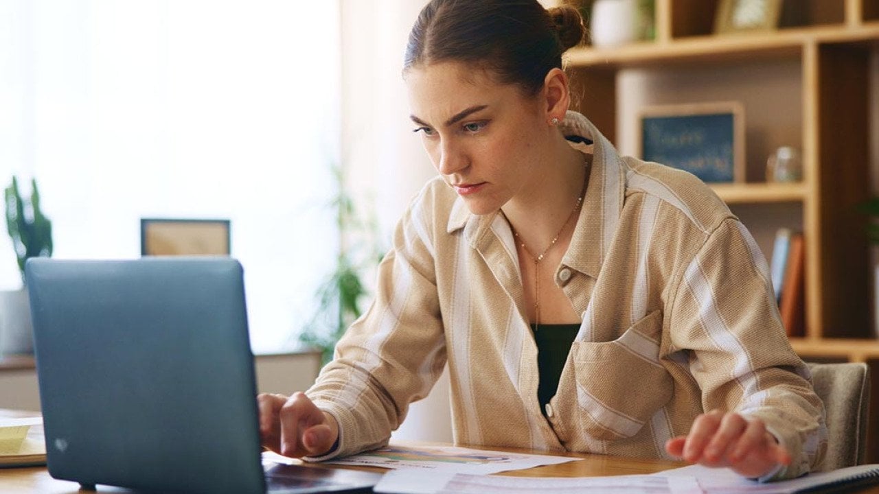 Determined-looking woman looking intently into laptop screen. She is sitting in an office with wooden bookshelves and a window behind her.