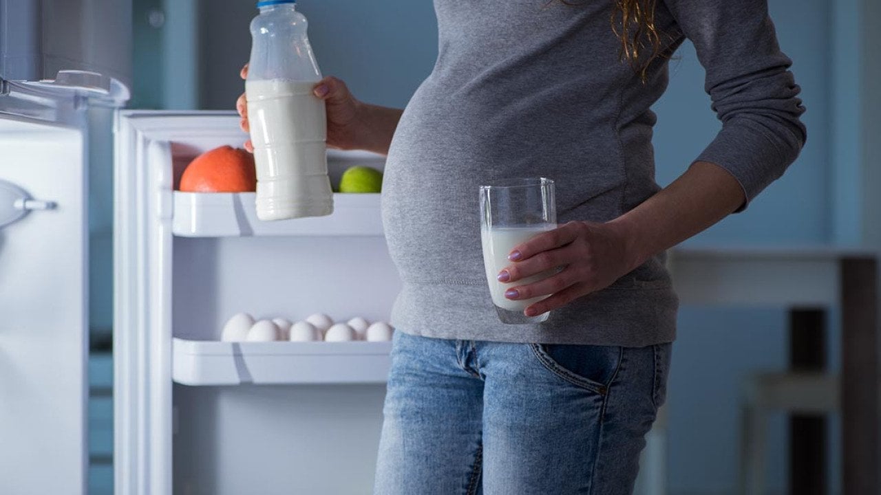 Pregnant woman, whose face is not shown, is illuminated by light of open fridge holding a glass in one hand and bottle of milk in the other.