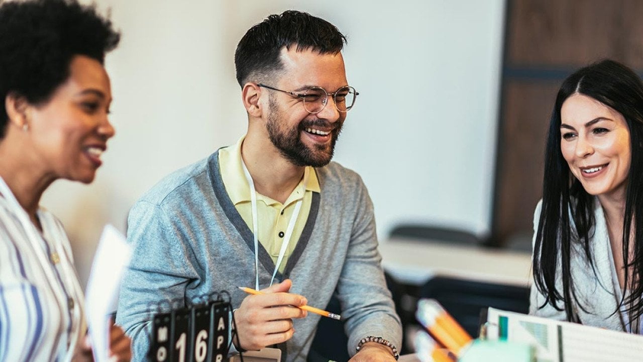Group of three sitting at a table at work. Man in the middle is laughing and holding a pencil and two women are on either side of him.