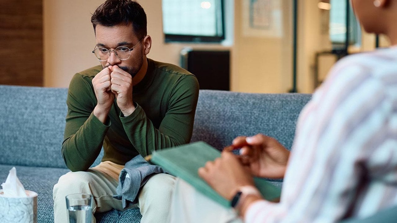 Man in a long sleeve green shirt wearing glasses sits on a blue couch breathing into his hands. A box of tissues is on the table and a therapist's back is in the foreground.