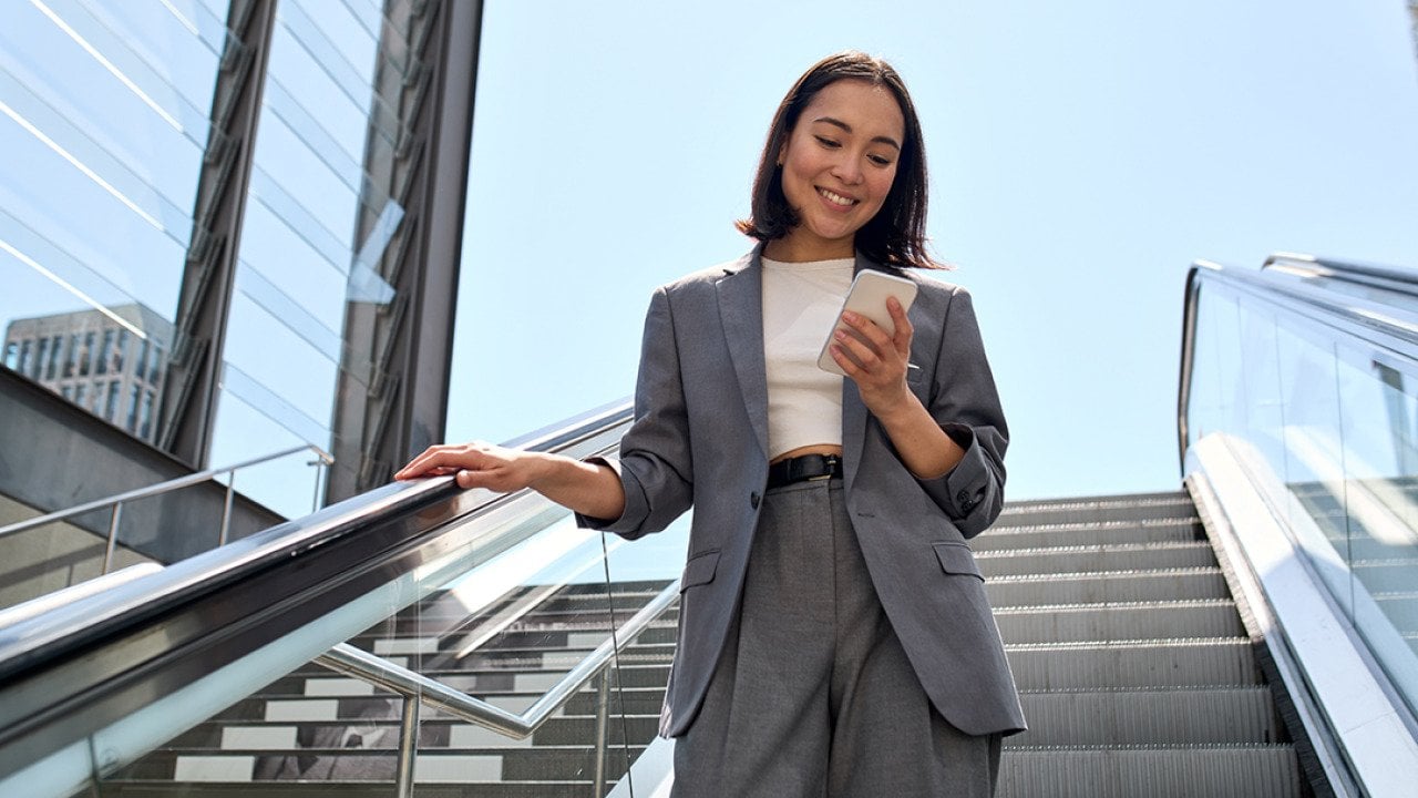 A young female marketing professional smiling at her phone while standing on an escalator.