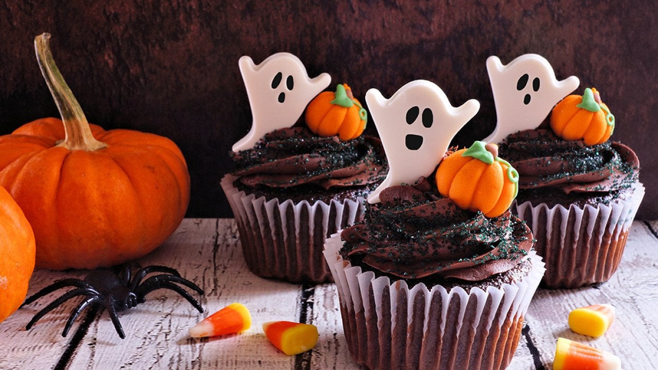 Chocolate cupcakes with ghost toppers beside Halloween pumpkins.